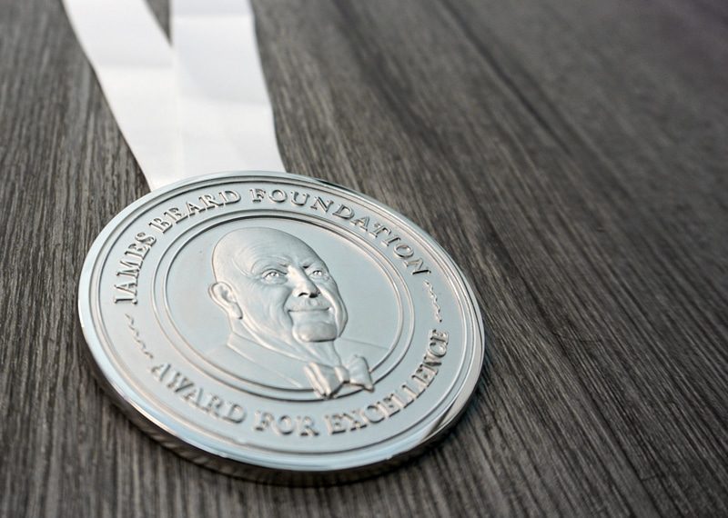James Beard Foundation medallion for chefs. Metal die stamped with text and image of face on the metal.
